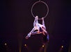 Aerial performer in a white bodysuit hangs upside down from a suspended hoop on a dimly lit stage with spotlights.