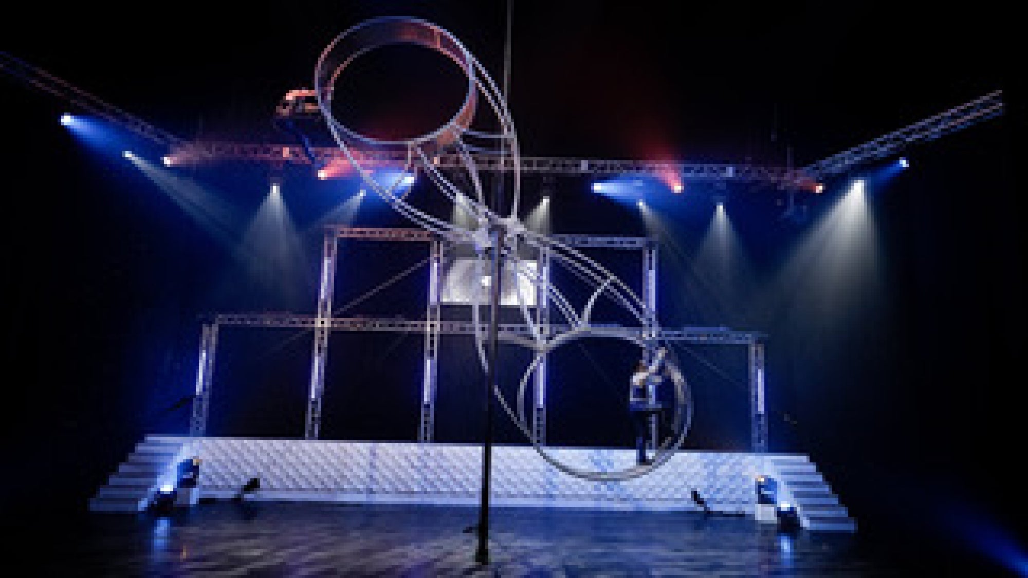 A person performs on a large spinning metal wheel apparatus on a stage with dramatic lighting and a truss background.