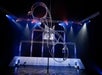 A person performs on a large spinning metal wheel apparatus on a stage with dramatic lighting and a truss background.
