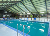 Indoor swimming pool with blue water, surrounded by blue lounge chairs and potted plants under a transparent roof.