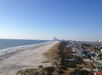 Aerial view of a coastal city with a long sandy beach, ocean waves, residential houses, and distant city skyline under a clear blue sky.