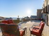 Sunlit patio with red cushioned chairs and a brick walkway overlooking the beach. A sign reads "Beach House" in the background, and the sun is low on the horizon.