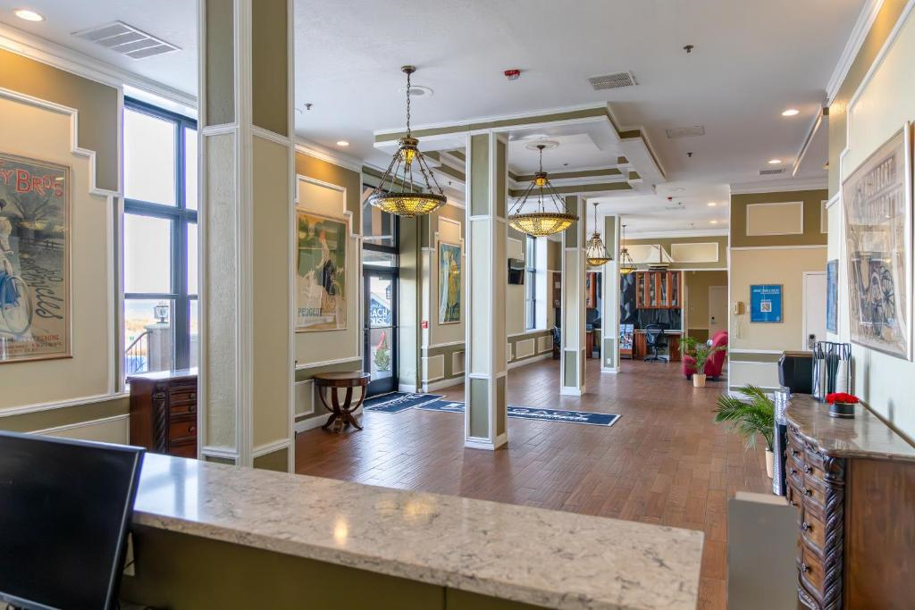 Hotel lobby with wooden flooring, a marble front desk, large windows, decorative chandeliers, and framed artwork on the walls. Red chairs and a small table are near the entrance.