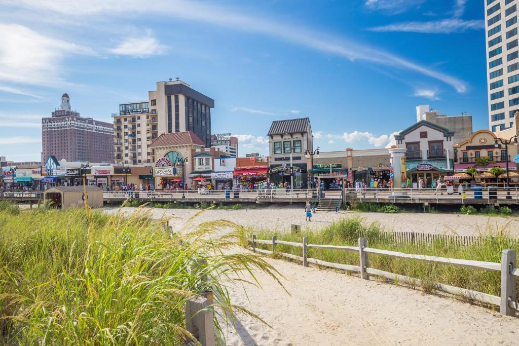 Beachfront boardwalk with people walking, surrounded by grass and sand; shops and tall buildings in the background under a blue sky.