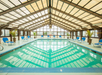 Indoor swimming pool with a sloped glass ceiling, surrounded by blue lounge chairs and potted plants, under soft lighting.