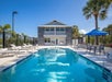 Outdoor pool area with lounge chairs, tables with umbrellas, and a two-story building in the background. Surrounded by palm trees and a black fence under a clear blue sky.