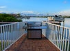 A stainless steel grill on a brick patio overlooking a calm waterfront with boats and a clear blue sky.