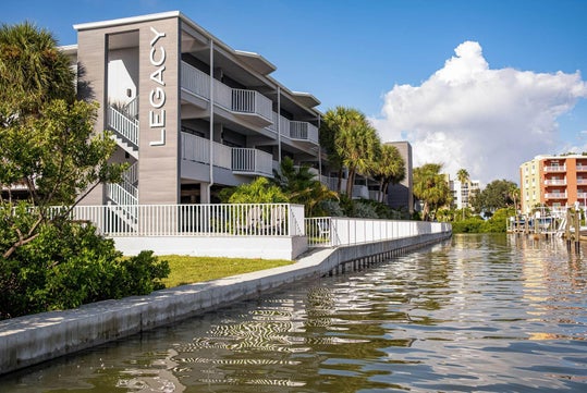 Three-story waterside apartment building with "Legacy" on the facade, surrounded by palm trees, against a blue sky with clouds.