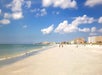 Sandy beach with scattered buildings on the right, a few people walking, and seagulls near the shore under a partly cloudy blue sky.
