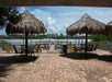 Outdoor waterfront patio with straw-roofed umbrellas, tables, and chairs on a paved terrace overlooking a body of water and distant buildings under a partly cloudy sky.