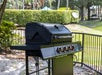 A black outdoor grill with multiple dials and a closed lid stands on a grassy area near a fence, surrounded by trees and bushes.