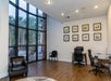 Office space with large windows, a brown armchair, two chairs, a desk with a computer, and framed pictures on the white wall. Outside, trees and patio furniture are visible.