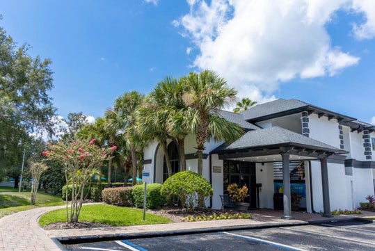 Modern building with palm trees, a pathway, and a small parking lot under a blue sky with clouds.