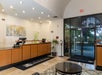 A hotel lobby with a reception desk, artwork on the wall, potted plants, and a glass exit door with outdoor view.