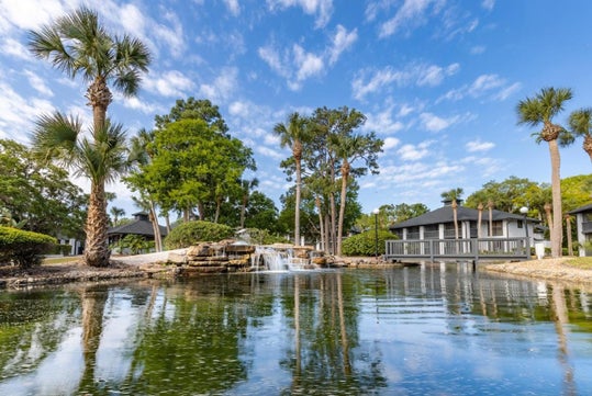 A serene pond with a small waterfall, surrounded by green trees and palm trees, reflects a white building with a dark roof under a blue sky with scattered clouds.