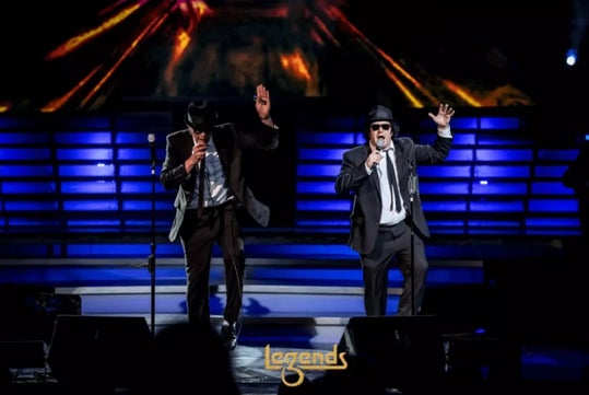 Two performers in black suits, hats, and sunglasses sing and gesture energetically on a brightly lit stage with "Legends" written at the front.