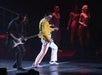 A performer in a yellow jacket and white pants sings on stage alongside a guitarist, with backup dancers standing in the background under stage lighting.