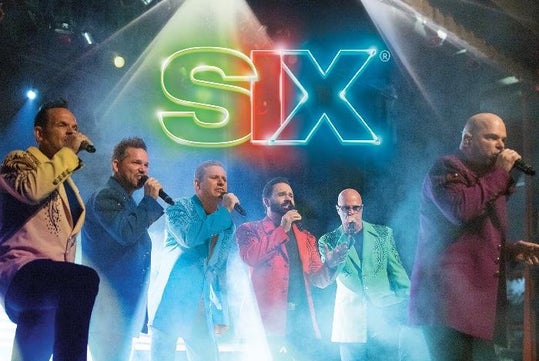 Six men in colorful suits sing into microphones on stage beneath a neon "SIX" sign, with stage lights in the background.