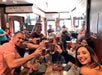 A group of people sit around a table in a bar, smiling and raising glasses filled with amber liquid in a toast. Other patrons are visible in the background.