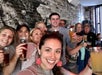 A group of people smiling and holding drinks in a bar with a brick wall backdrop.