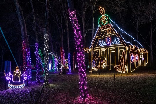 A nighttime outdoor scene with trees wrapped in colorful holiday lights and a large illuminated house-shaped decoration in the background.