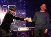 A magician in a velvet jacket performs a card trick with a smiling man on stage, under starry and digital backdrop.