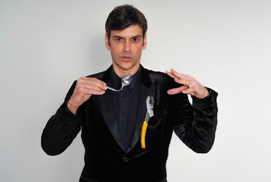 Man in a black suit holds a bent spoon with pliers in his jacket pocket.