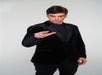 A person in a black suit and shirt makes a gesture with one hand, standing against a plain background.