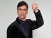 A man in a black suit raises his hand with fingers pinched together against a plain background, looking forward.