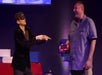 A blindfolded performer points while a smiling man in a blue shirt holds a large red digital clock on a stage with a red table and blue props in the background.