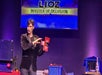 A man in a black suit performs a magic trick on stage in front of a sign that reads "Lioz Master of Delusion." Various props are on the table next to him.