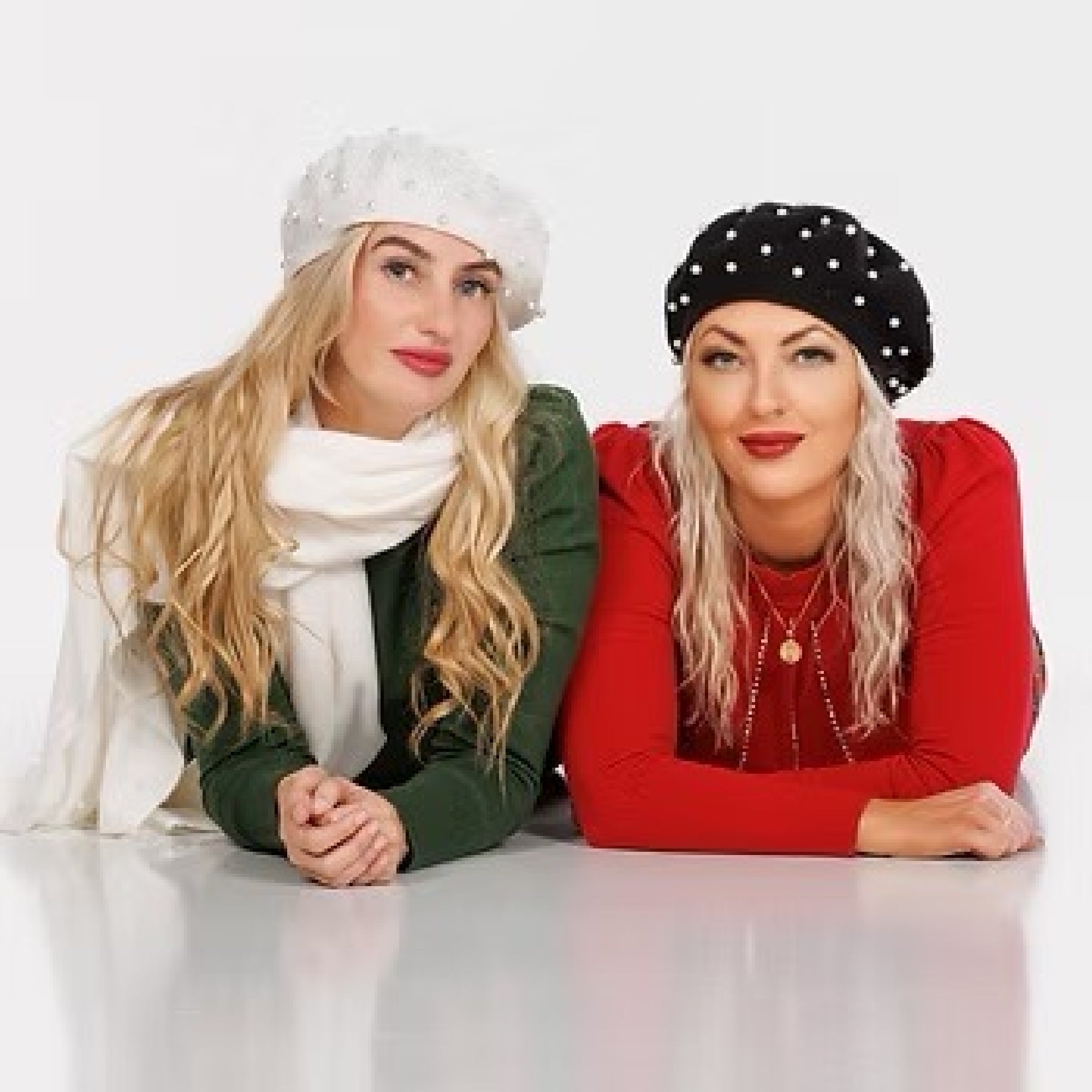 Two women wearing berets and long-sleeve tops lie side by side on the floor, facing forward. One wears green and white, the other wears red and black. Both have long, blonde hair.