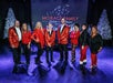 Eight people wearing festive red and black outfits stand onstage in front of a sign that reads "Morace Family Listen to the Music Branson," with white Christmas trees in the background.