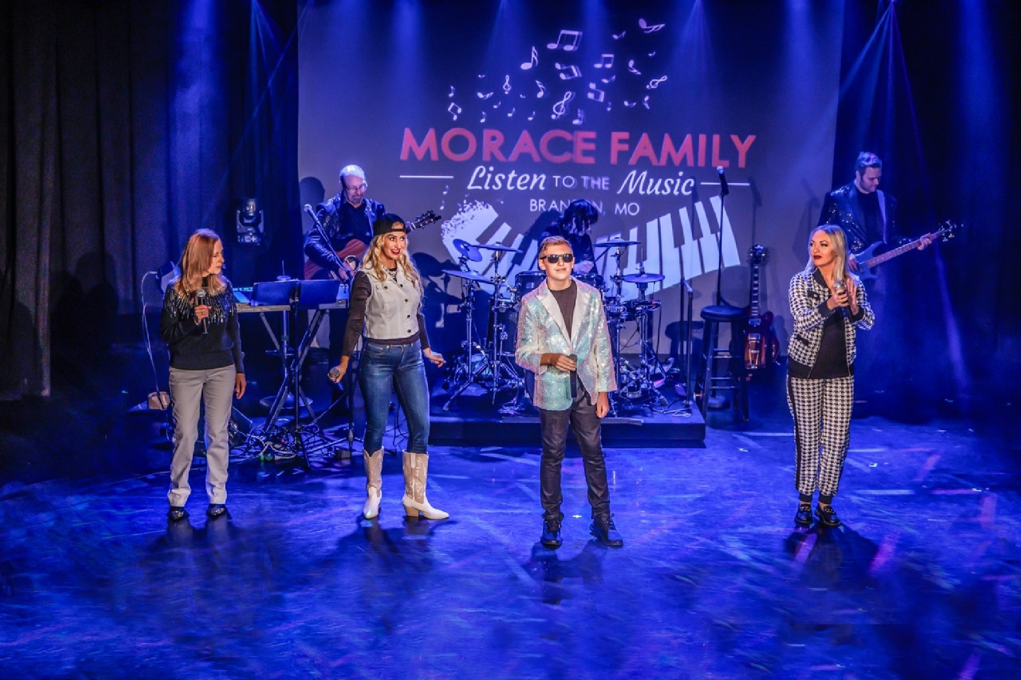 Five performers stand on stage with musical instruments in the background under a sign that reads, "Morace Family Listen to the Music Branson MO." Blue and purple stage lighting is used.