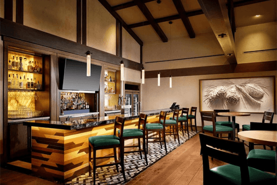 Modern bar with wooden accents, illuminated shelves of bottles, a flat screen TV, and green cushioned chairs. Tables with chairs are arranged around the room under pendant lights.