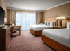 Hotel room with two neatly made beds, a desk with a chair, a flat-screen TV, and a window with sheer curtains. Room features warm tones and wooden furniture.