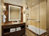 Modern bathroom with glass-enclosed bathtub, brown marbled countertop, large mirror, and wall-mounted lights. Towels are stored below the sink.