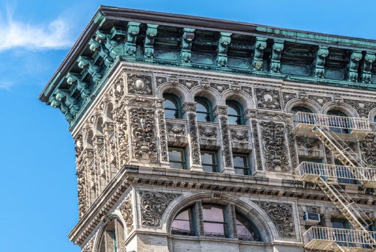 An ornate, multi-story building corner with detailed classical architecture and a green patina cornice against a clear blue sky.