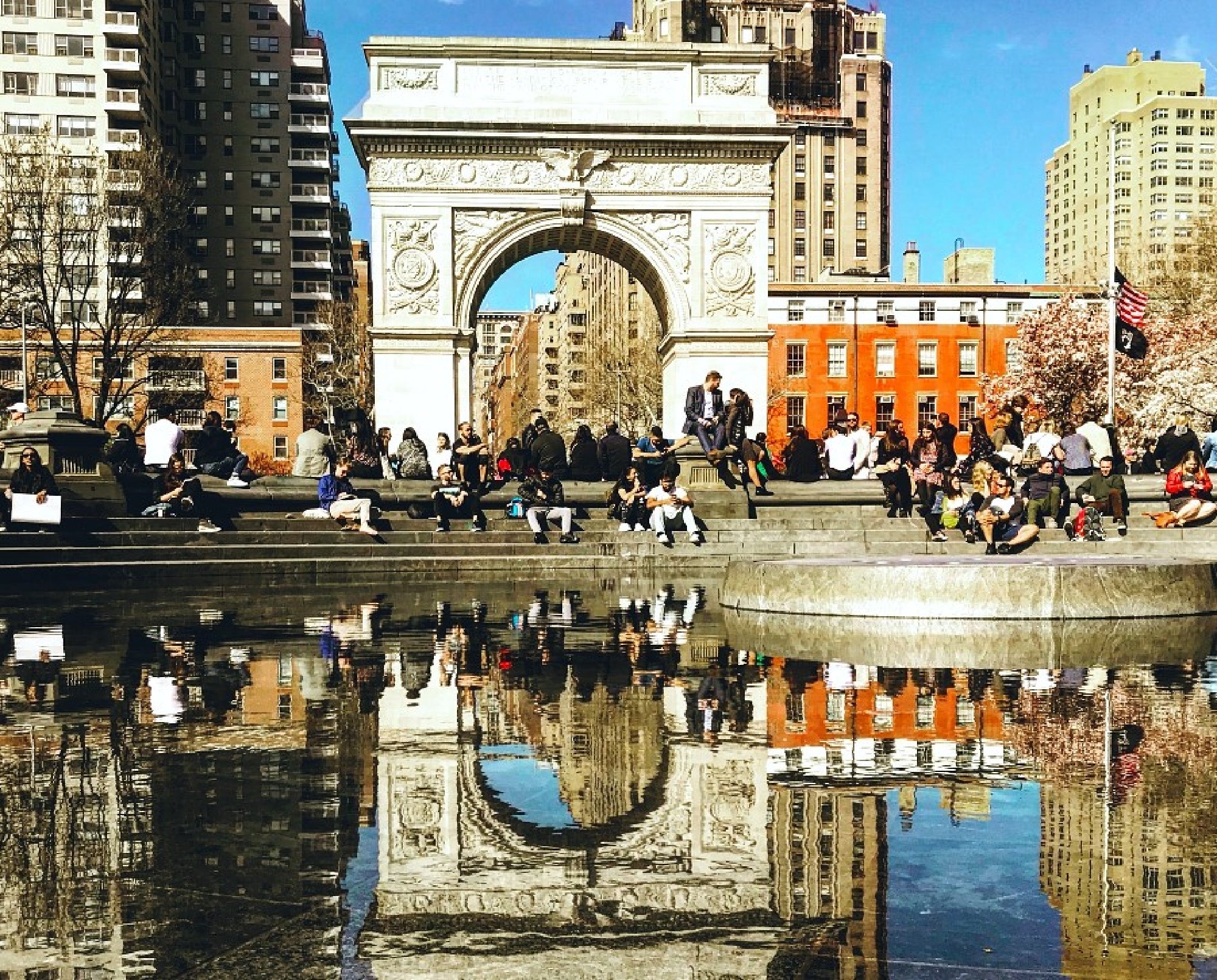 People are gathered around a fountain in front of a large arch in a city park, with several buildings in the background and reflections visible in the water.