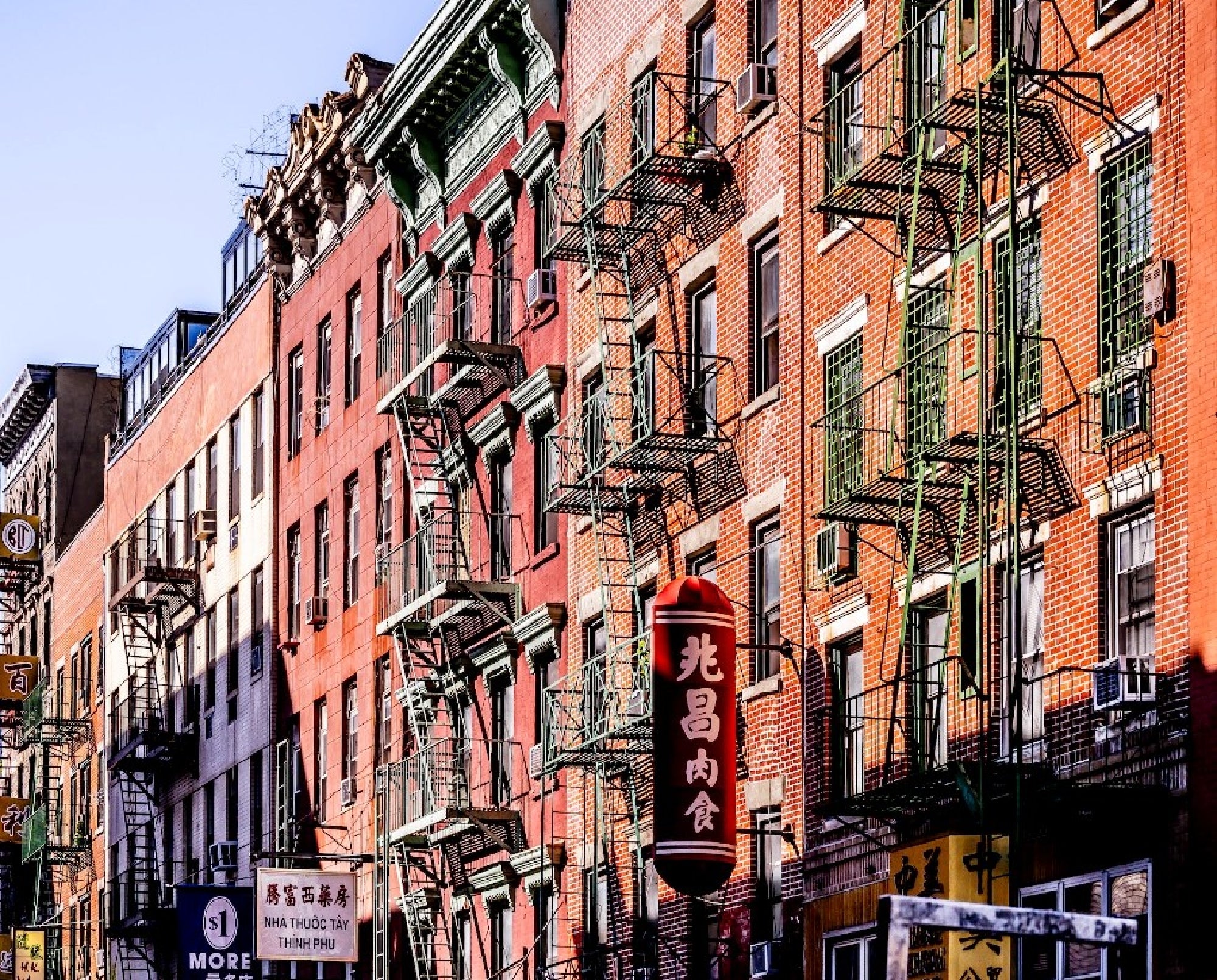 A row of urban buildings with fire escapes and various signs, including a prominent red sign with Chinese characters in front, set against a clear sky.