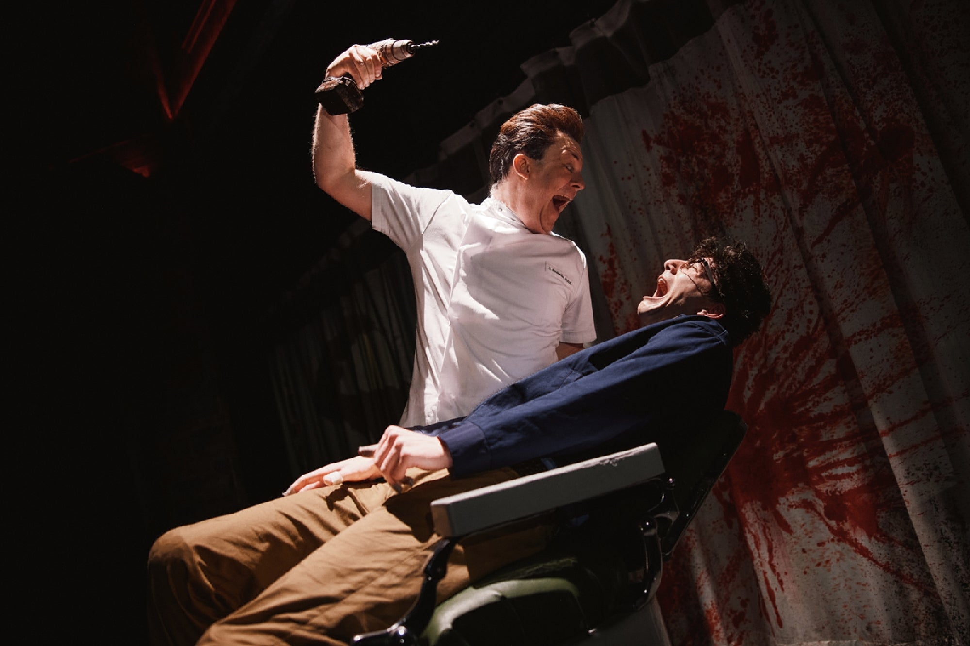 A person in a dentist's uniform holds a dental drill menacingly over another person sitting in a chair, both with exaggerated expressions; bloody splatters cover the background curtain.