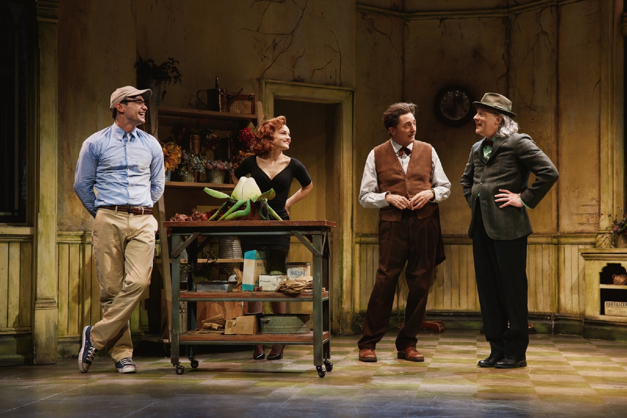 Four actors stand in a vintage kitchen set, smiling and conversing around a cart with food items, in a scene from a theatrical play.