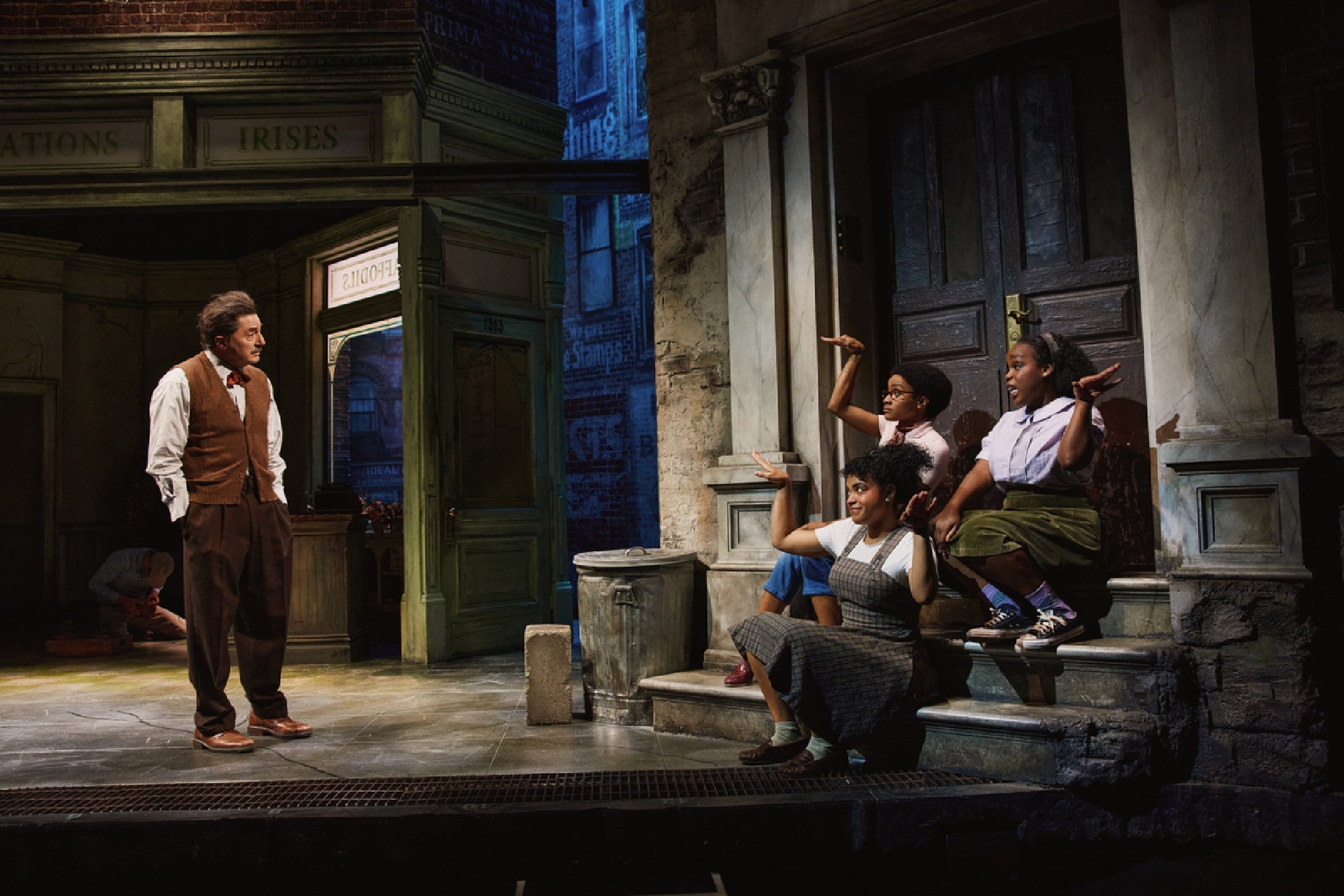 A man stands onstage facing three women seated on steps in front of a building facade, in a theatrical stage set with urban scenery.