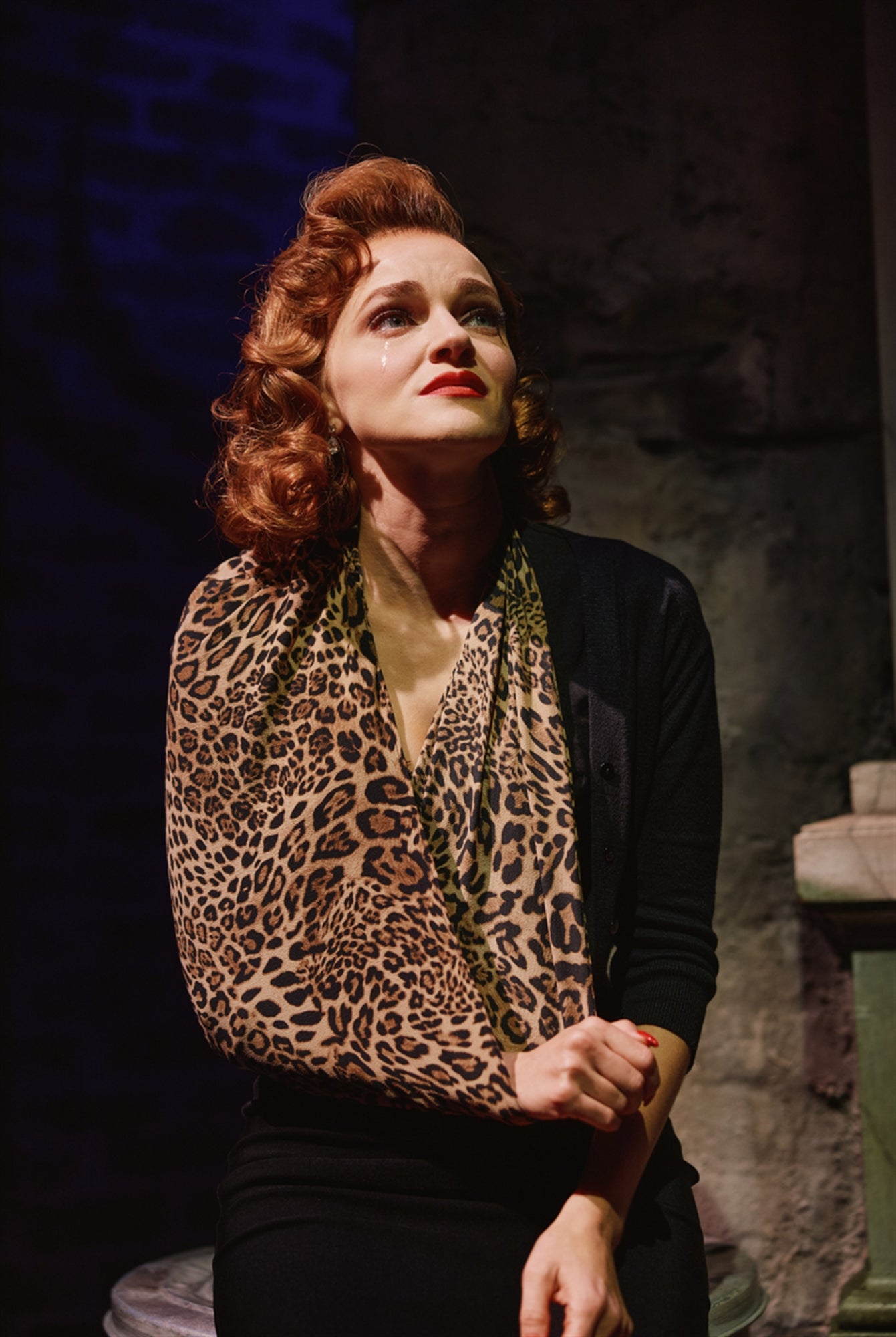 A woman with red hair and red lipstick, wearing a leopard print scarf and black outfit, sits on stage with a tear on her cheek, looking upward under dramatic lighting.