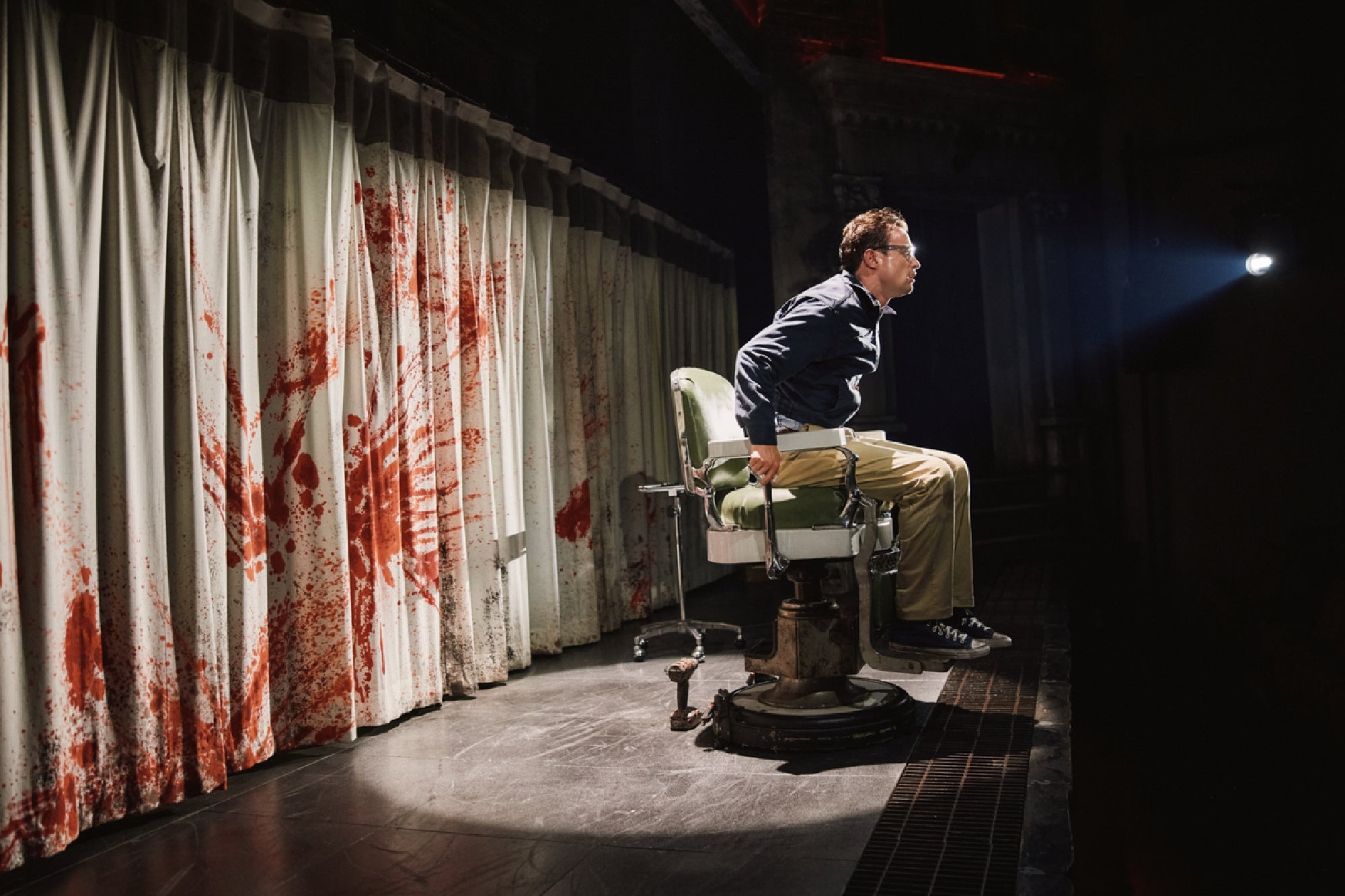 A man in glasses sits on the edge of a vintage barber chair. Behind him, white curtains are stained with large, smeared red marks resembling blood. A spotlight shines on the scene.