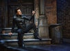 A man in a black leather jacket and boots sits on steps by a brick building, pointing with one hand, next to a metal trash can.