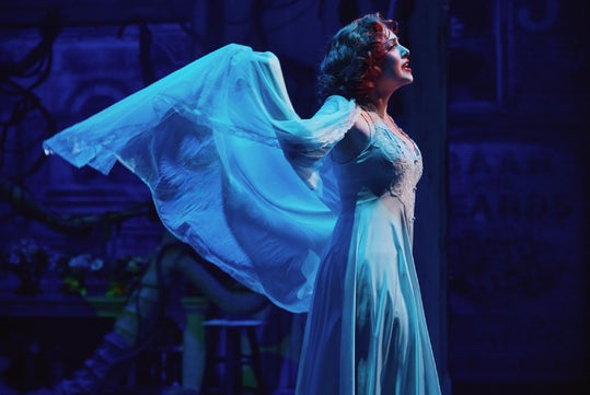 A woman in a long, flowing dress stands on stage with her arms raised, holding sheer fabric, under dramatic blue lighting.