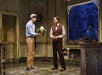 Two men stand in an old-fashioned flower shop; one holds a large green and white plant, while the other listens, framed by a blue-tinted window displaying a city scene outside.
