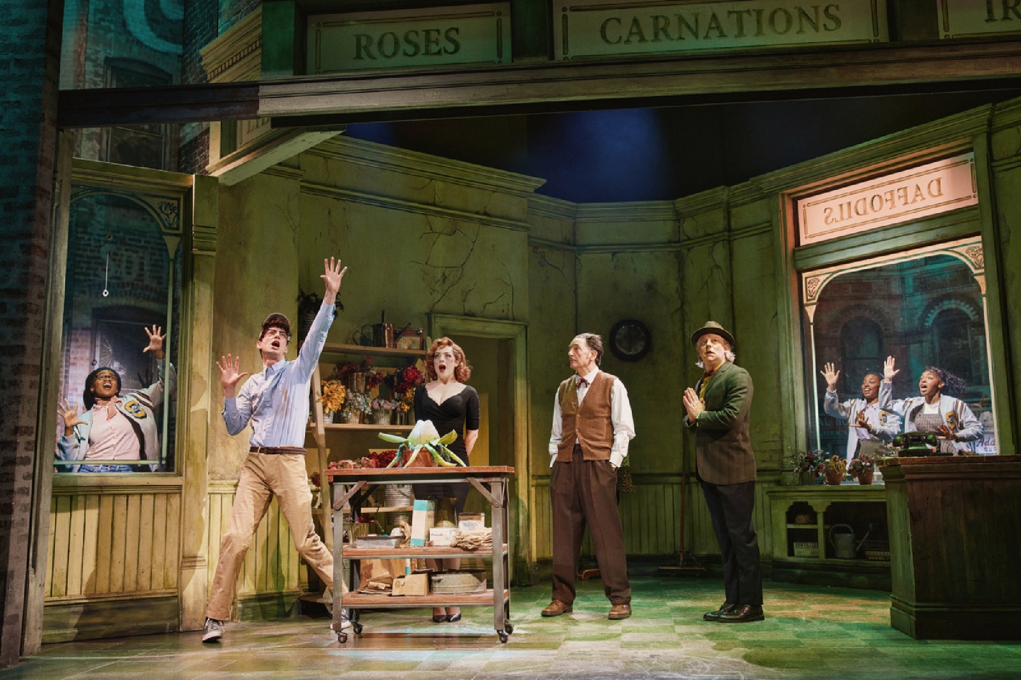 Actors perform on a stage set designed as a flower shop, with dramatic poses and expressions, surrounded by labeled sections for roses, carnations, and daffodils.