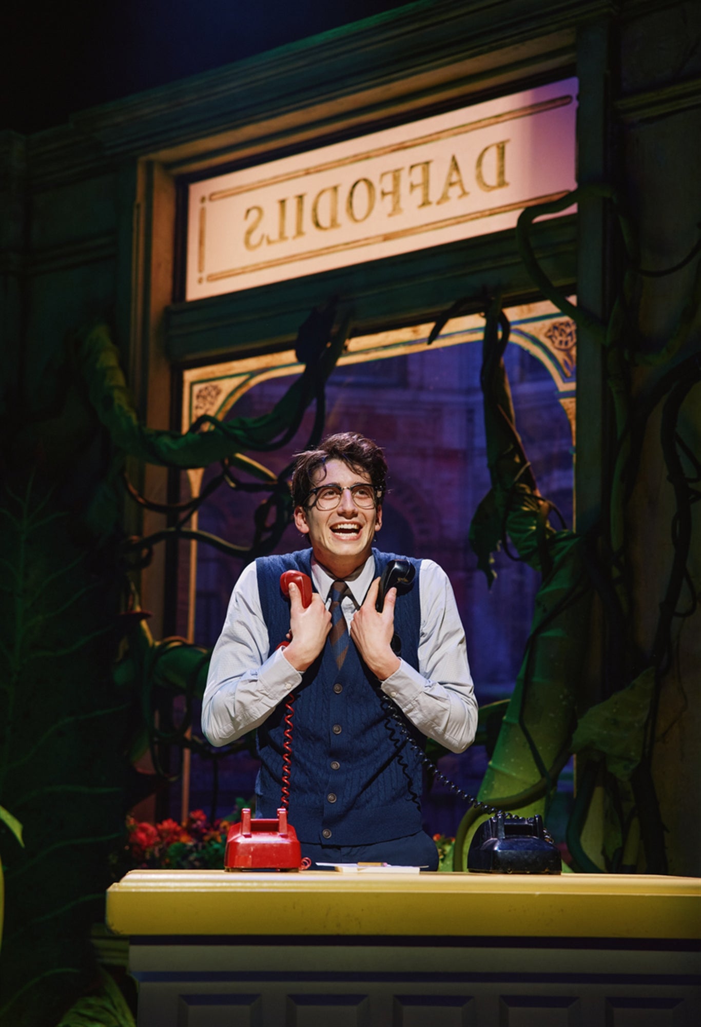 A man in glasses and a vest stands at a counter, holding two telephones with a smile. A sign reading "DAFFODILS" is visible in the background, along with large green plants.